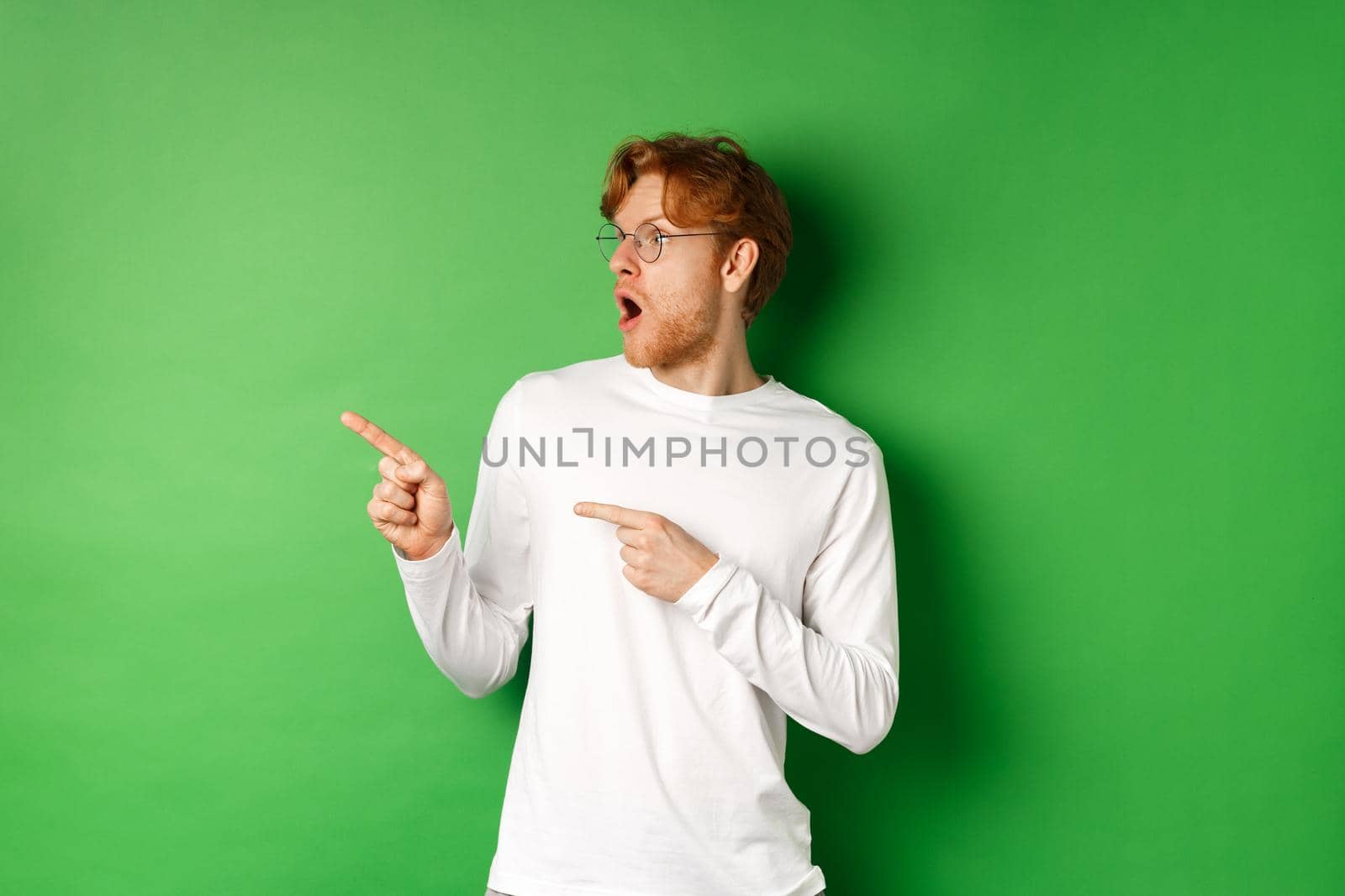 Impressed redhead man in glasses checking out promo, pointing and looking left with amazed face, standing in long-sleeve t-shirt over green background.