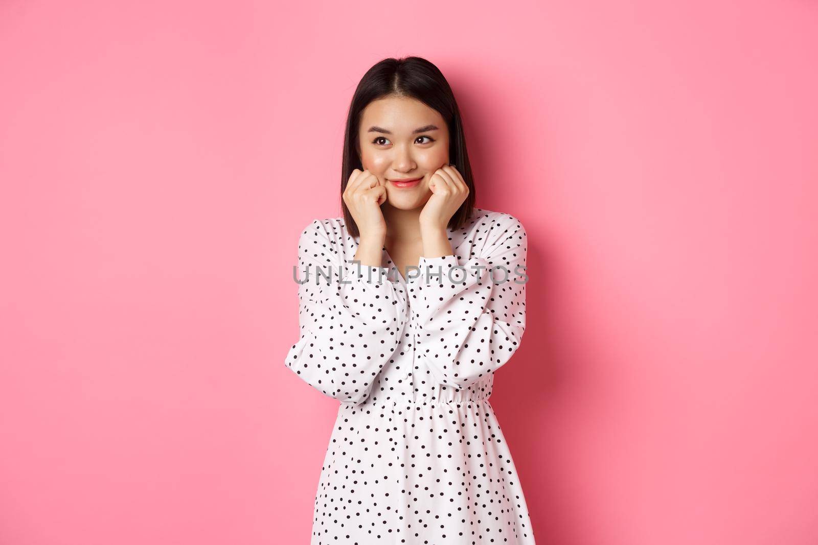 Cute and shy asian girl blushing, touching cheeks and looking left at copy space silly, standing against pink background by Benzoix