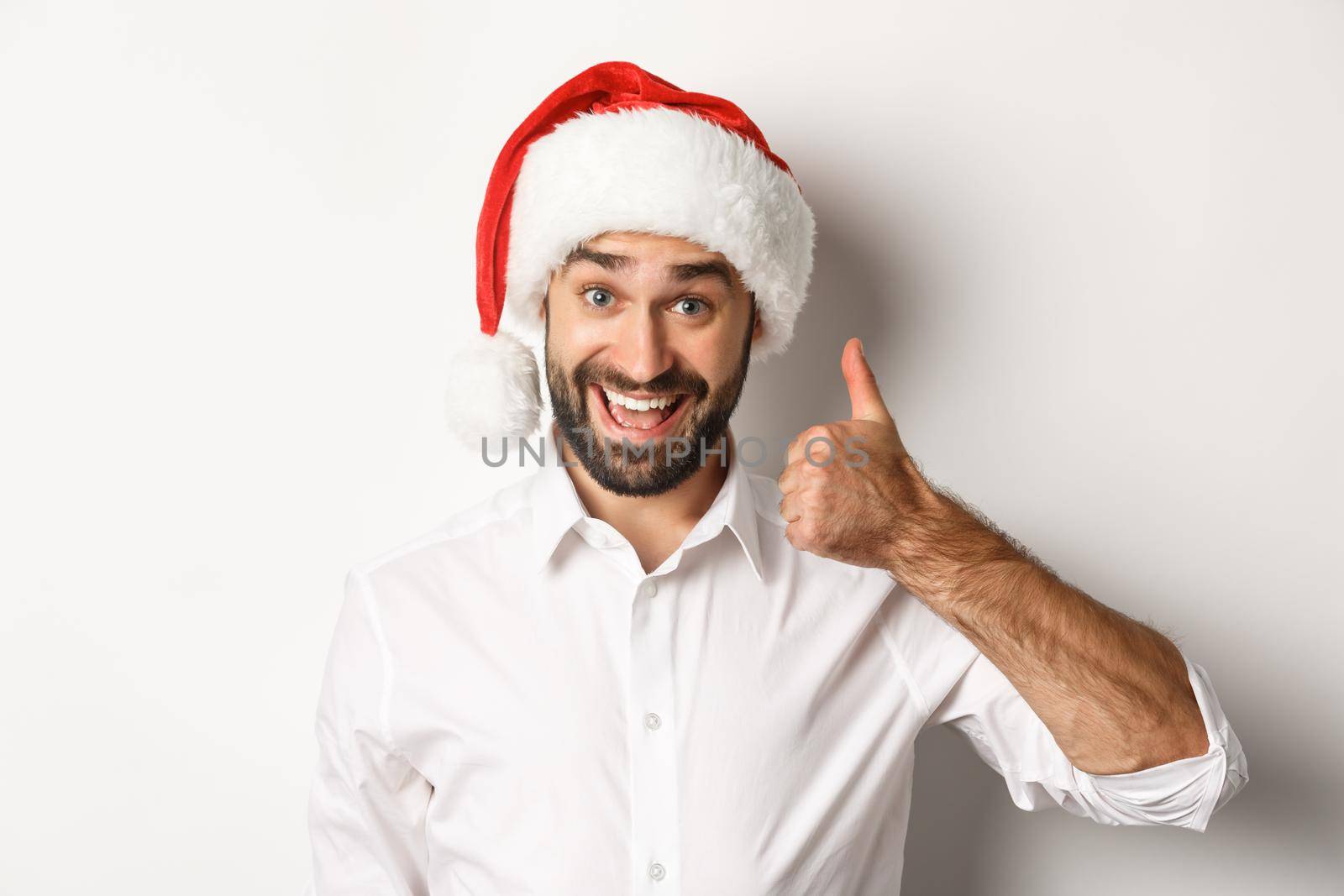 Party, winter holidays and celebration concept. Close-up of satisfied bearded man in santa hat showing thumb up, approve and like something good, white background by Benzoix