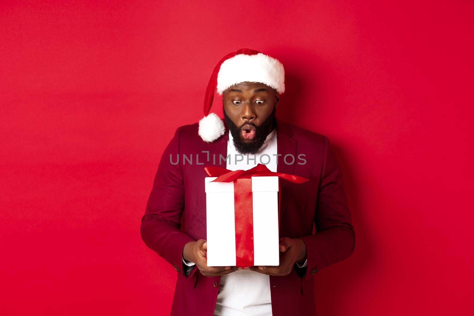 Christmas, New Year and shopping concept. Surprised Black man staring at xmas present, saying wow amazed, receiving holiday gift, red background.