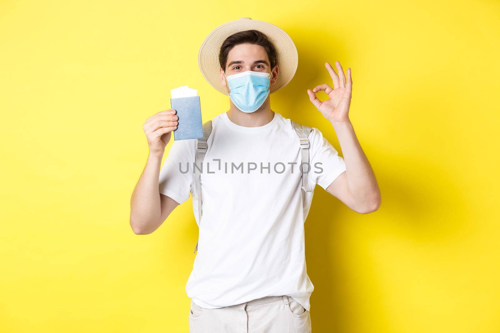 Concept of covid-19, tourism and pandemic. Happy male tourist in medical mask showing passport, going on vacation during coronavirus, make ok sign, yellow background.