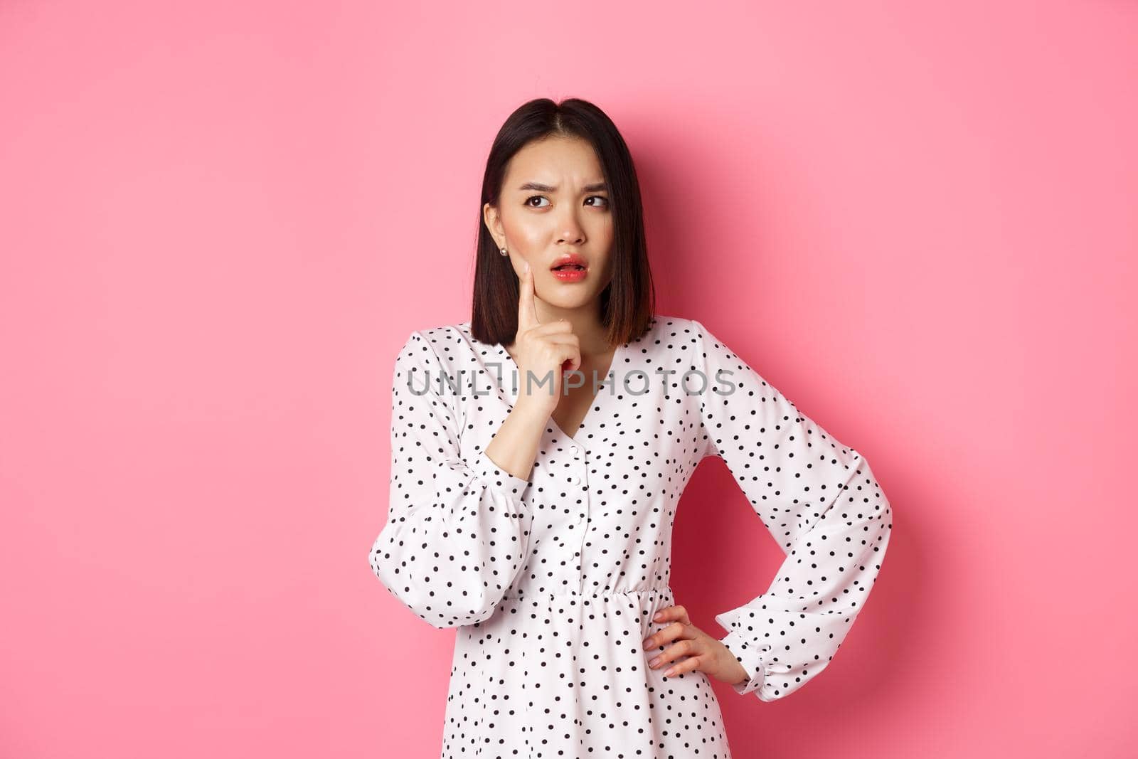 Thoughtful asian woman making assumption, looking up and thinking, deciding something, standing in dress over pink background by Benzoix