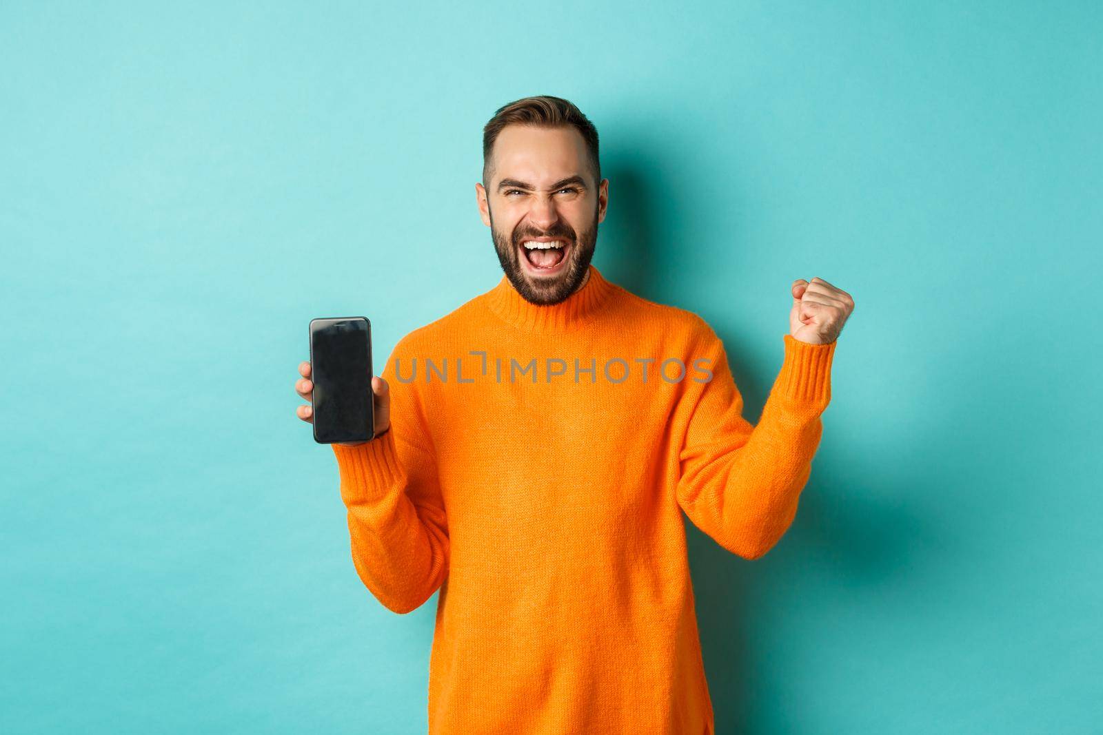 Image of happy young man winning and showing mobile phone screen, rejoicing and celebrating victory, fist pump with satisfaction, standing over light blue background.