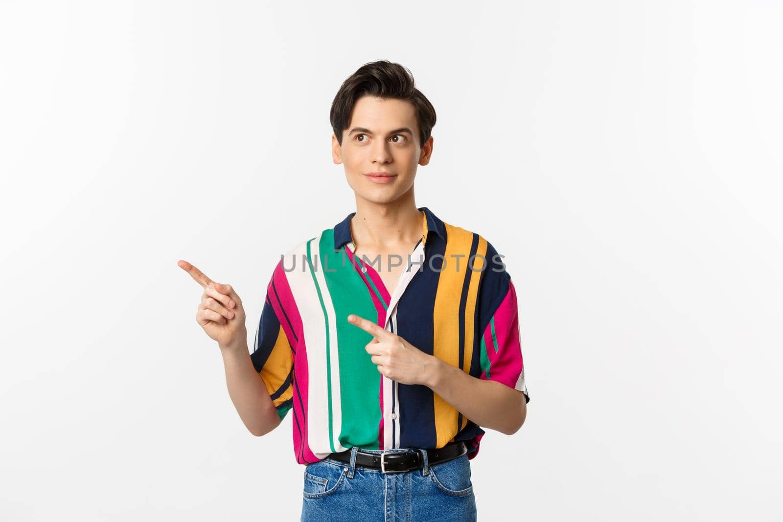 Young handsome queer man pointing, looking left at logo, smiling pleased, standing over white background.