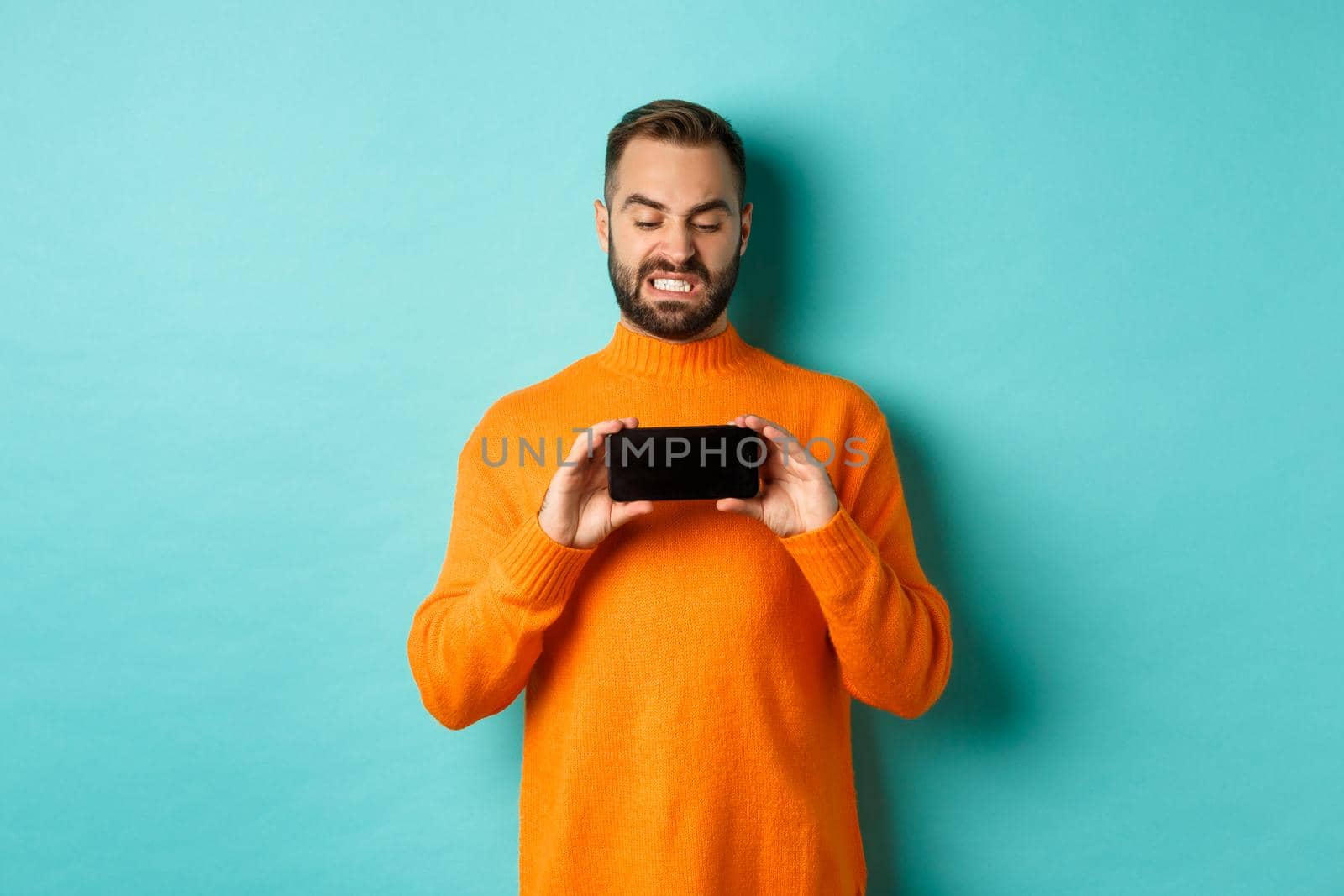 Image of man cringe at something on mobile screen, staring disgusted at display, showing smartphone, standing over light blue background.