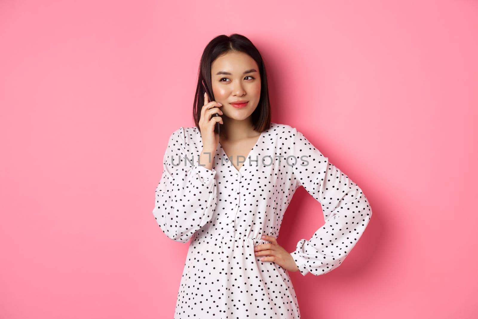 Attractive korean woman making a phone call, holding smartphone near ear and smiling, standing over pink background.