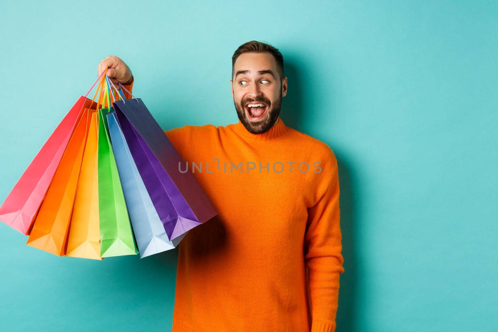 Happy young man going shopping, holding bags and looking excited, standing in orange sweater over blue background.