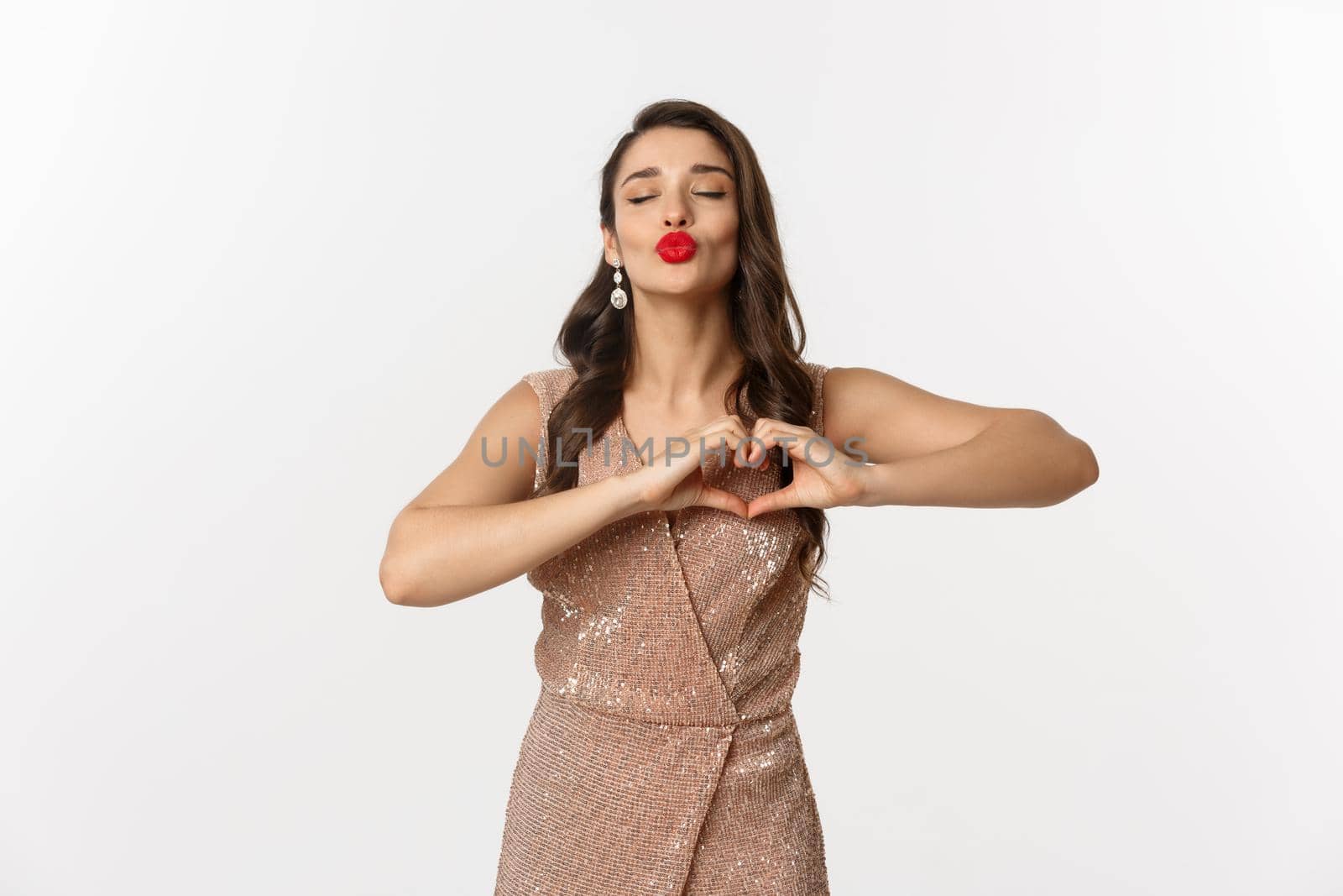Celebration and party concept. Beautiful young woman in stylish dress, red lips, showing heart sign and waiting for kiss, standing over white background by Benzoix