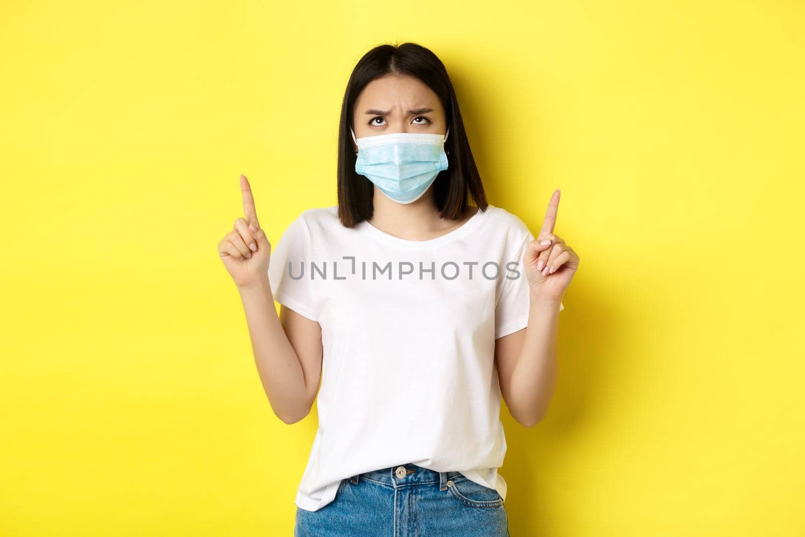 Covid-19, pandemic and social distancing concept. Disappointed asian girl in medical mask, frowning upset and pointing fingers up at logo, standing over yellow background.