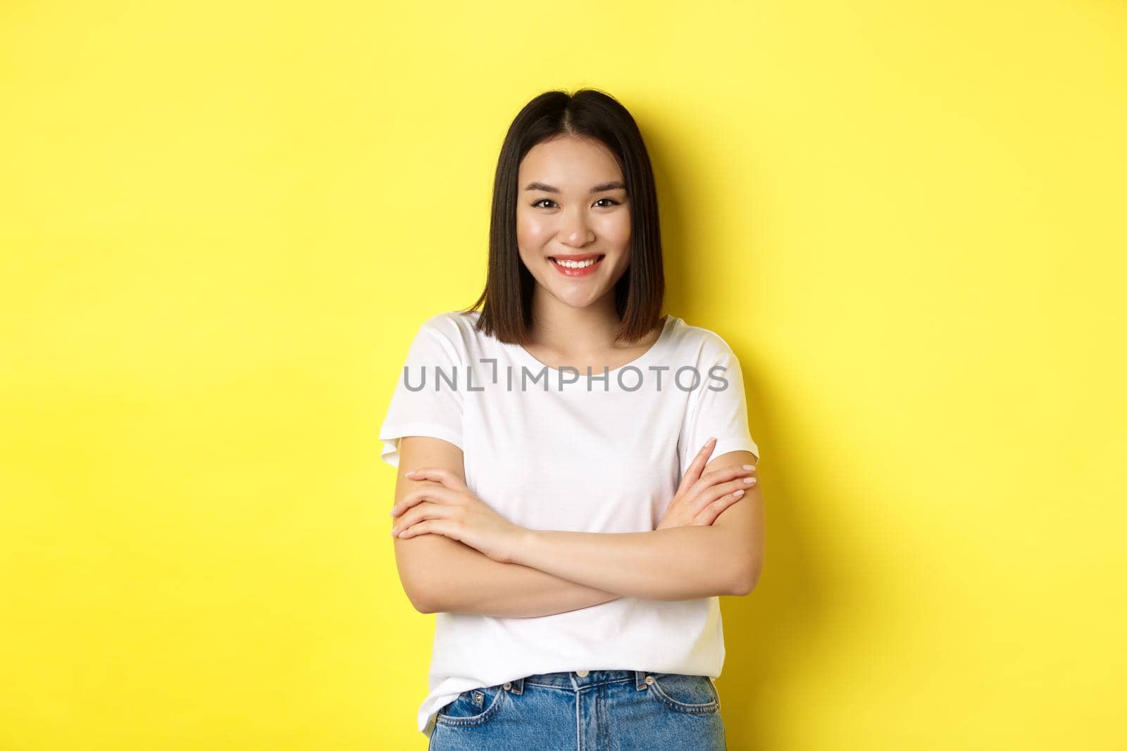 Confident and stylish asian woman cross arms on chest and smiling, standing over yellow background by Benzoix