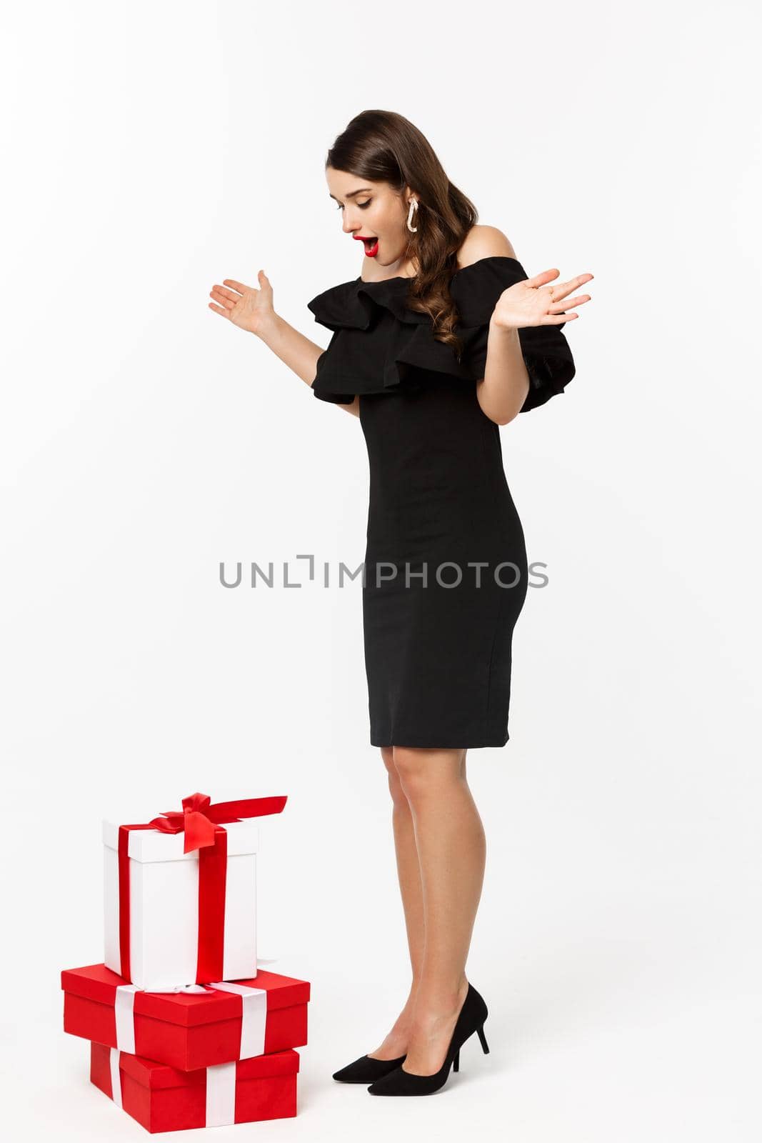 Full length view of woman in elegant dress and red lips, looking surprised, receive gifts on christmas holidays, standing with presents over white background.