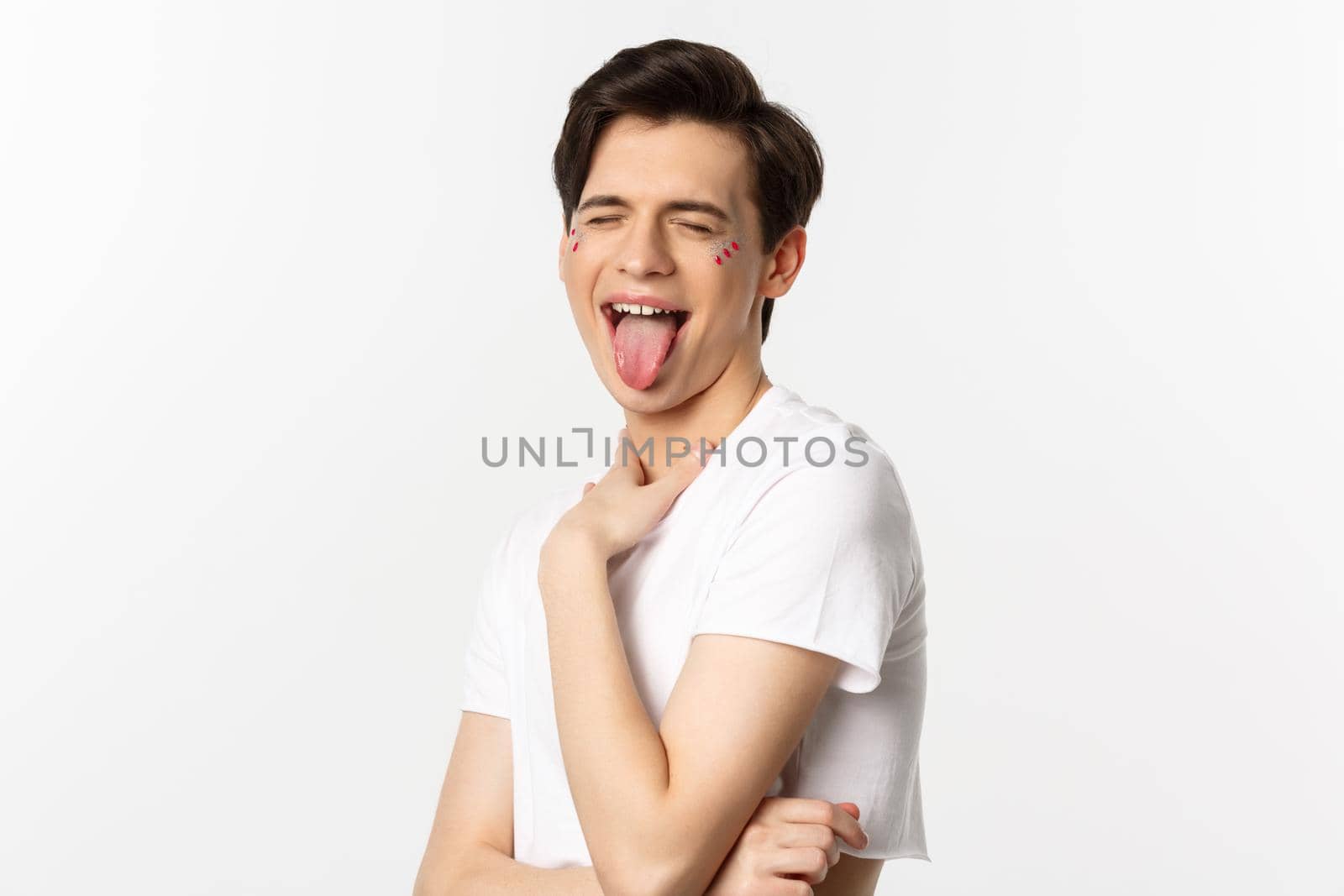 People, lgbtq and beauty concept. Close-up of carefree gay man with glitter on face, showing tongue and making funny face, standing over white background.