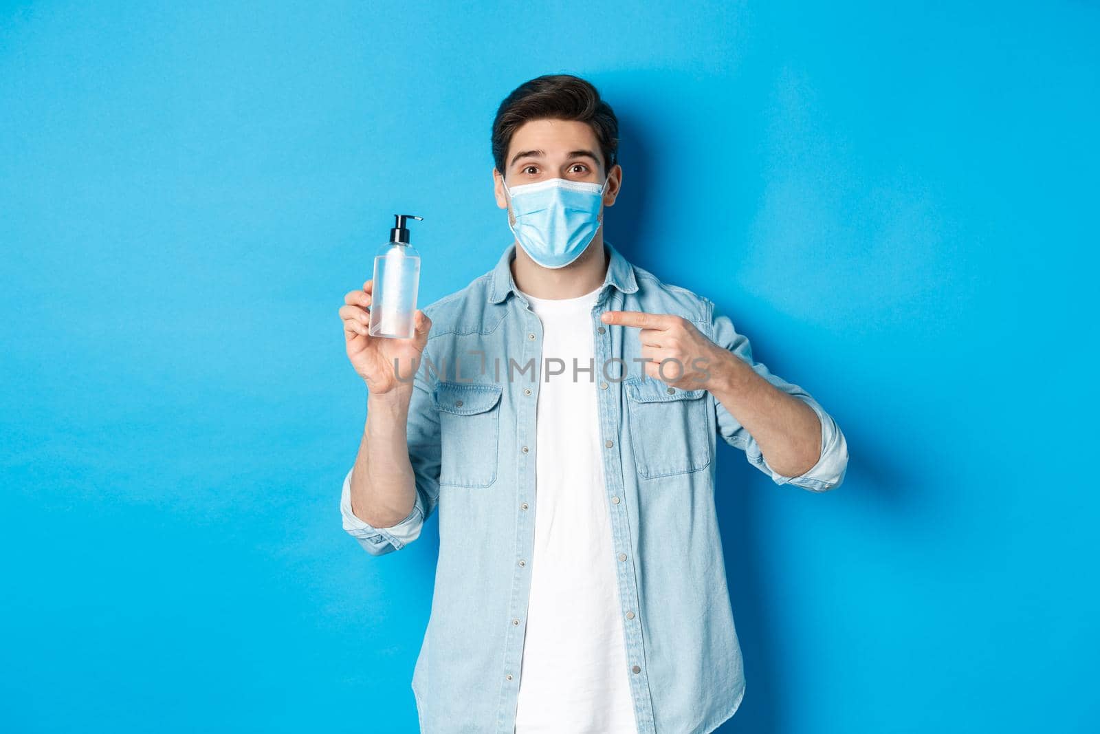 Concept of covid-19, pandemic and social distancing. Handsome guy in medical mask advice to use hand sanitizer, pointing at antiseptic, standing over blue background.