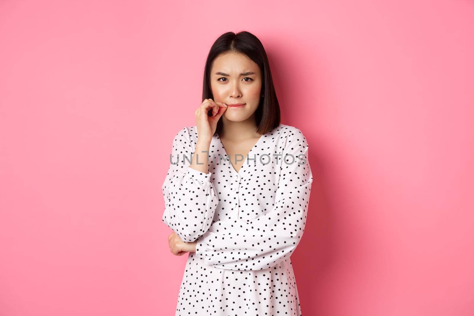 Brunette asian woman in dress looking displeased, frowning and zipping mouth, seal lips with promise, standing over pink background.