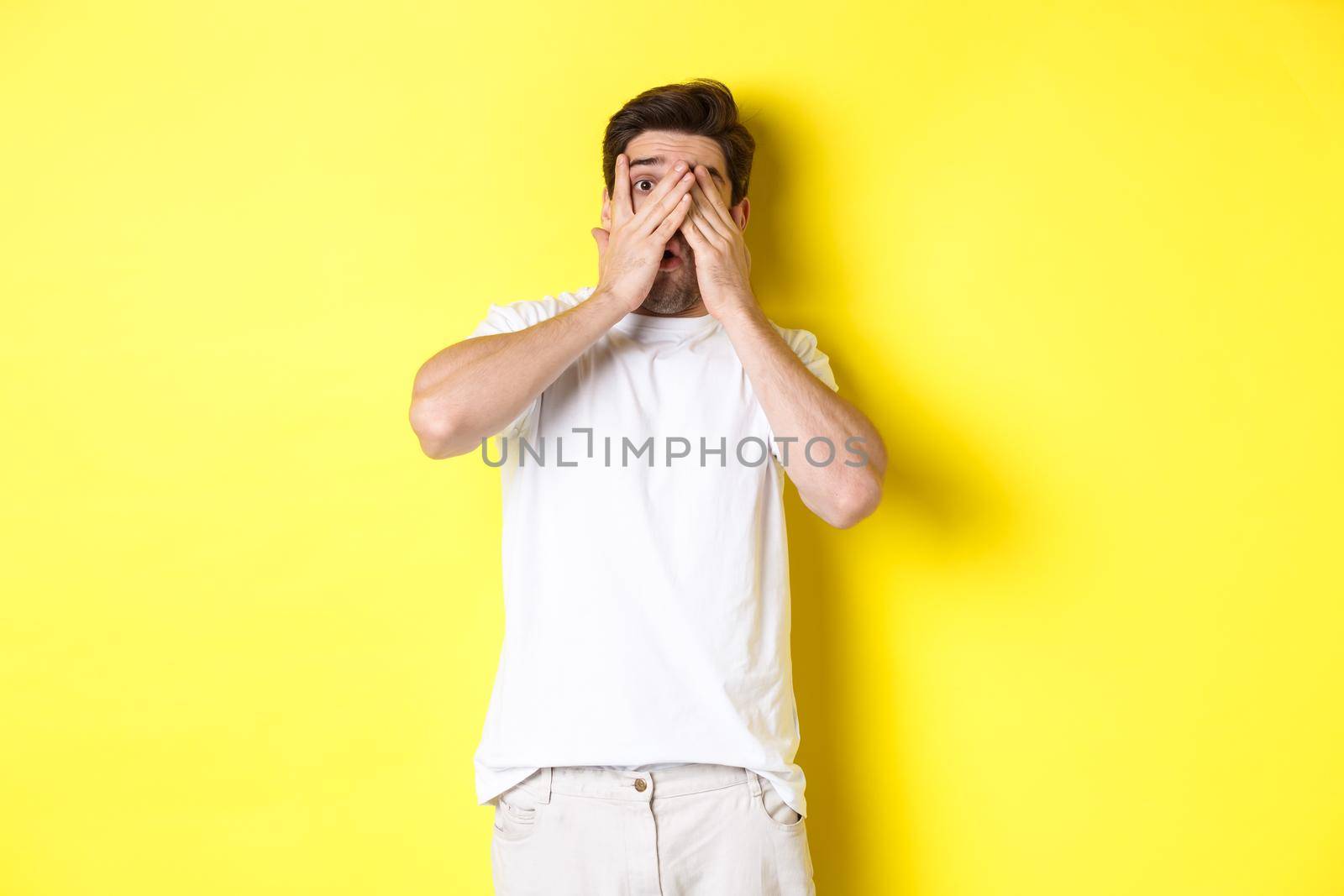 Embarrassed guy shut eyes but peeking through fingers at something awkward, standing over yellow background.