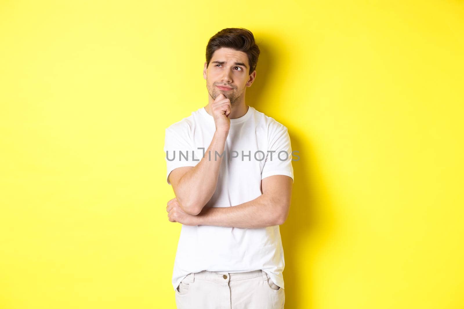 Portrait of young male model thinking, looking at upper left corner and making choice, standing near copy space, yellow background.