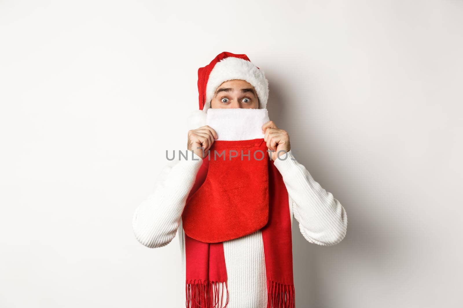 Winter holidays and shopping concept. Cheerful caucasian guy in Santa hat hiding face behind Christmas sock with gifts, standing over white background.