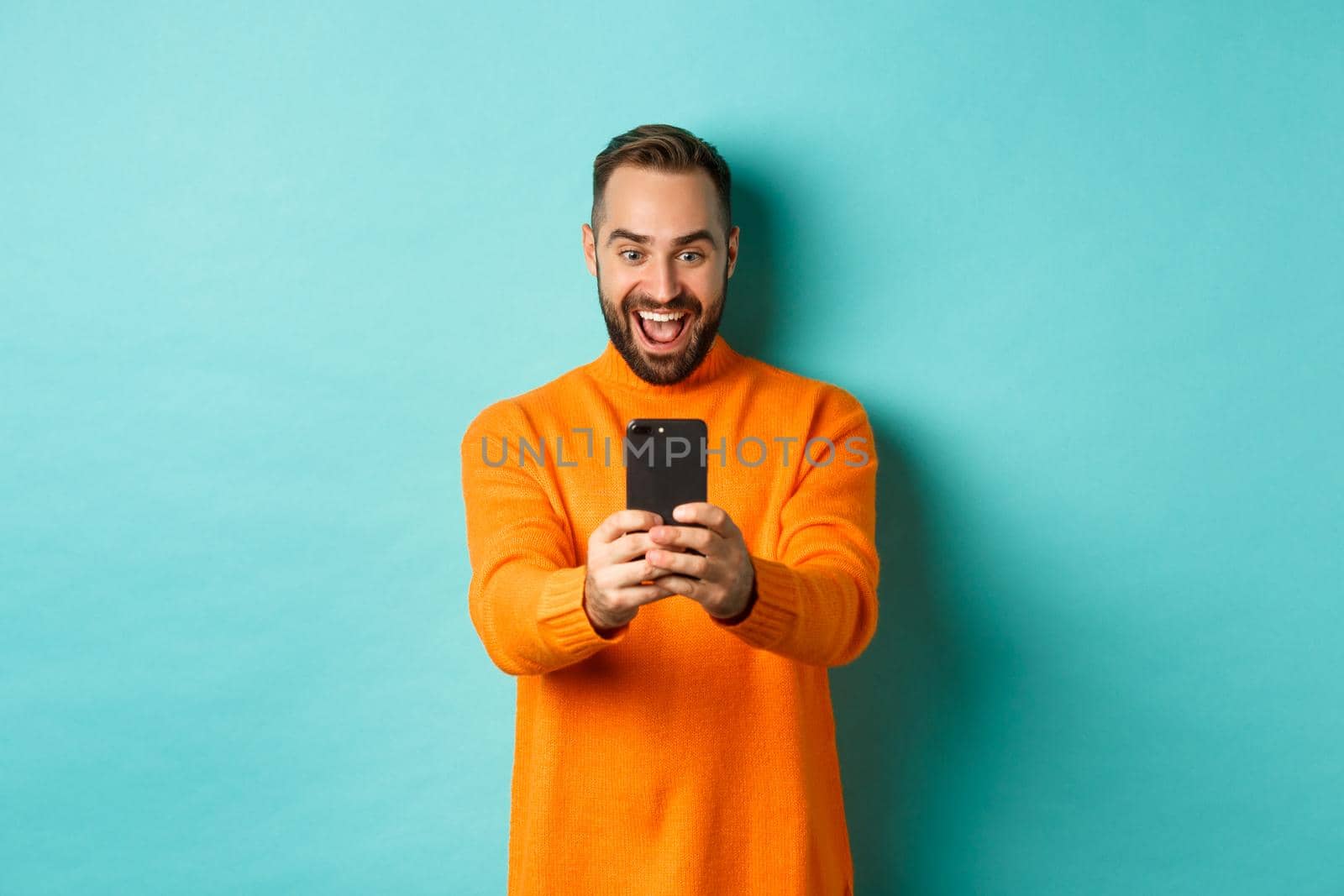 Image of man reading awesome message on mobile phone, looking at smartphone screen with amazement, standing over light blue background by Benzoix