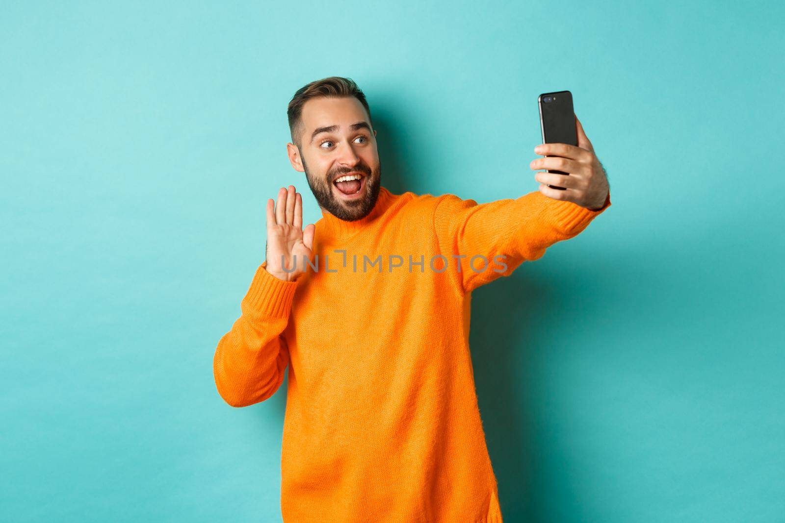 Happy young man video calling, talking online with mobile phone, saying hello to smartphone camera and waving hand friendly, standing over light blue background.