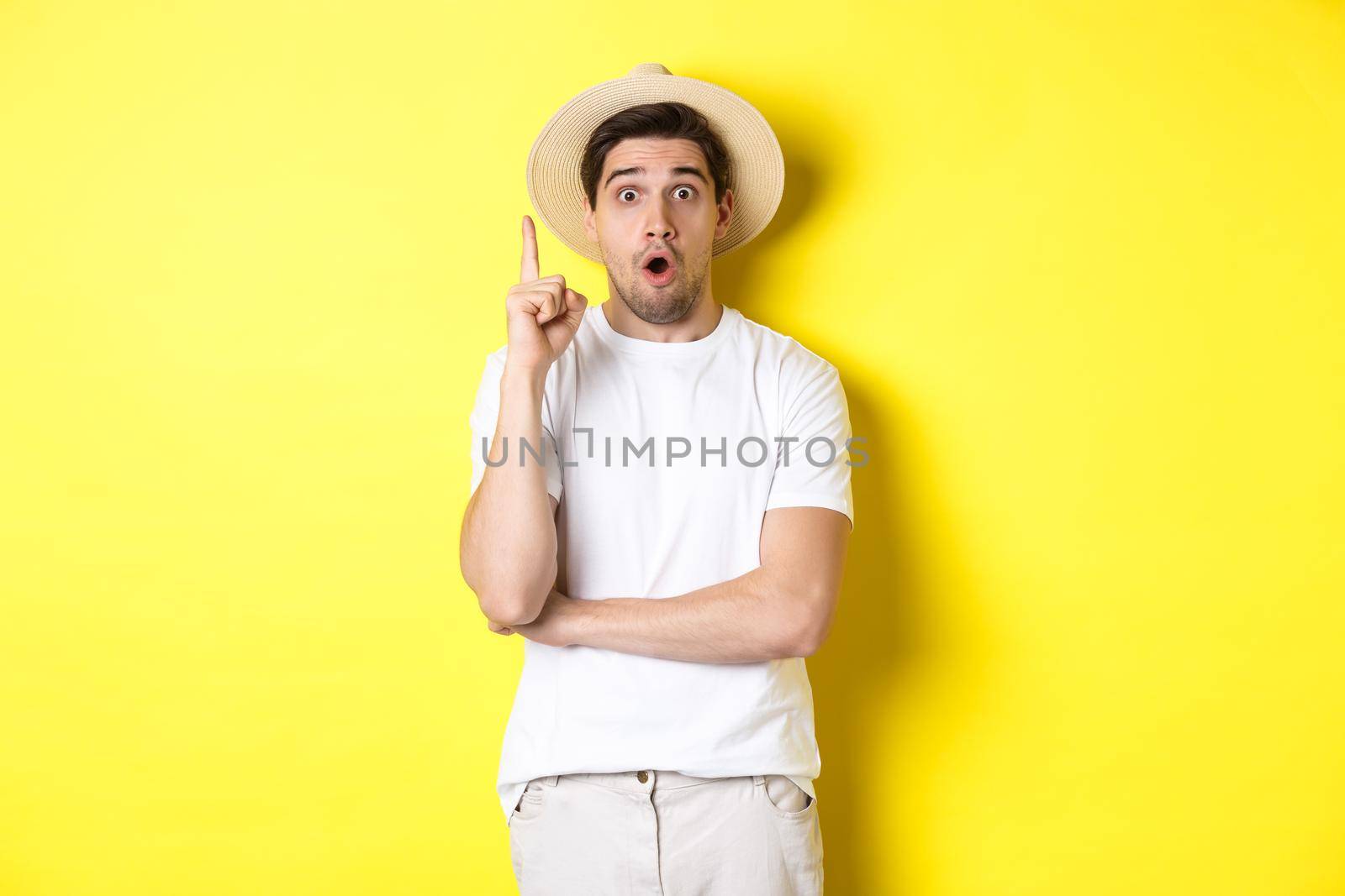 Portrait of young man in straw hat having an idea, raising finger eureka sign, making suggestion, standing over yellow background.