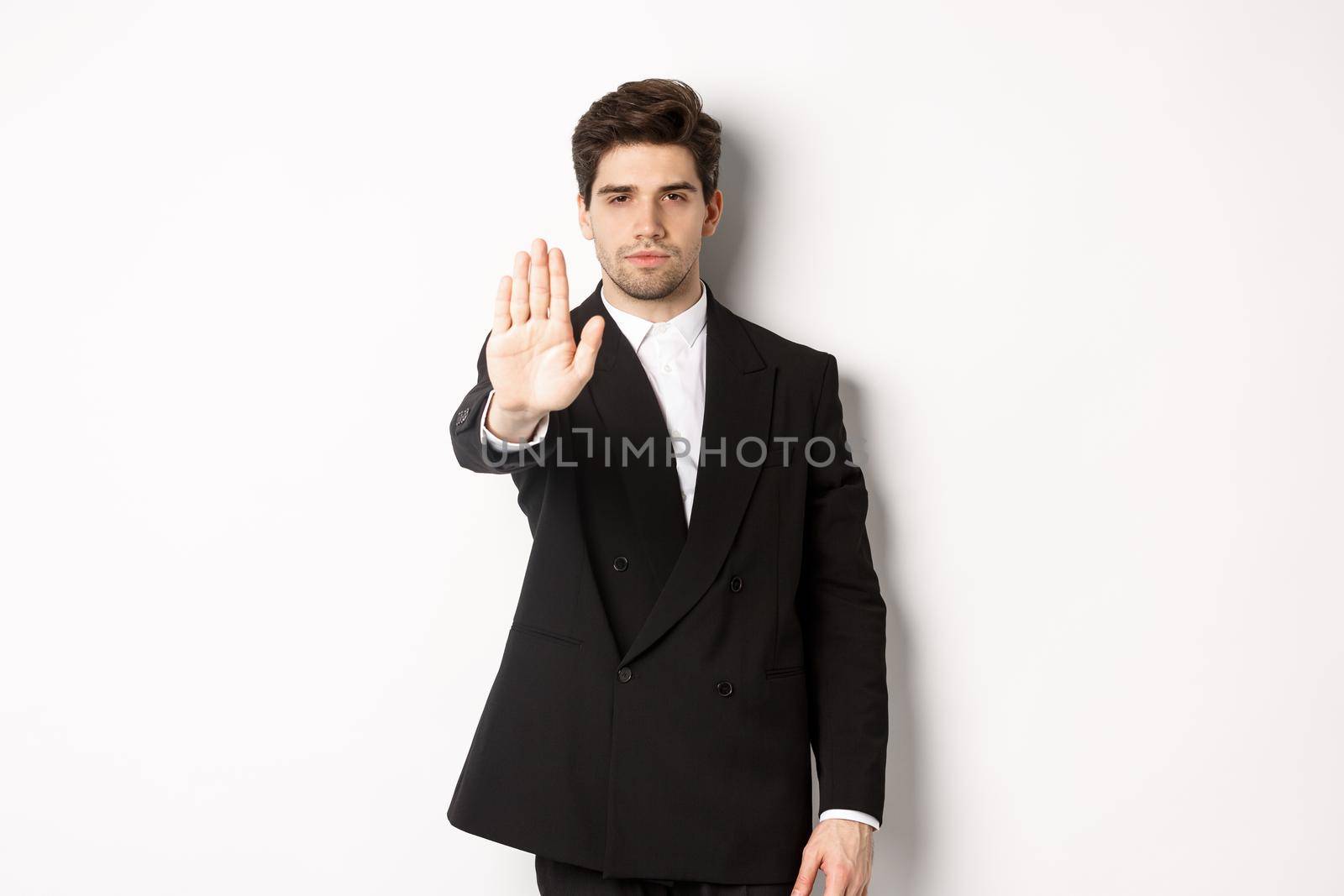 Portrait of serious handsome man in formal suit, extending hand to stop you, prohibit action, forbid and disagree with something, standing against white background.