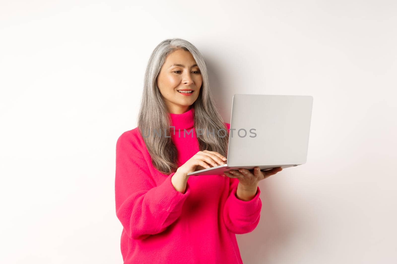 Senior asian woman working freelance, using laptop and smiling, standing over white background.