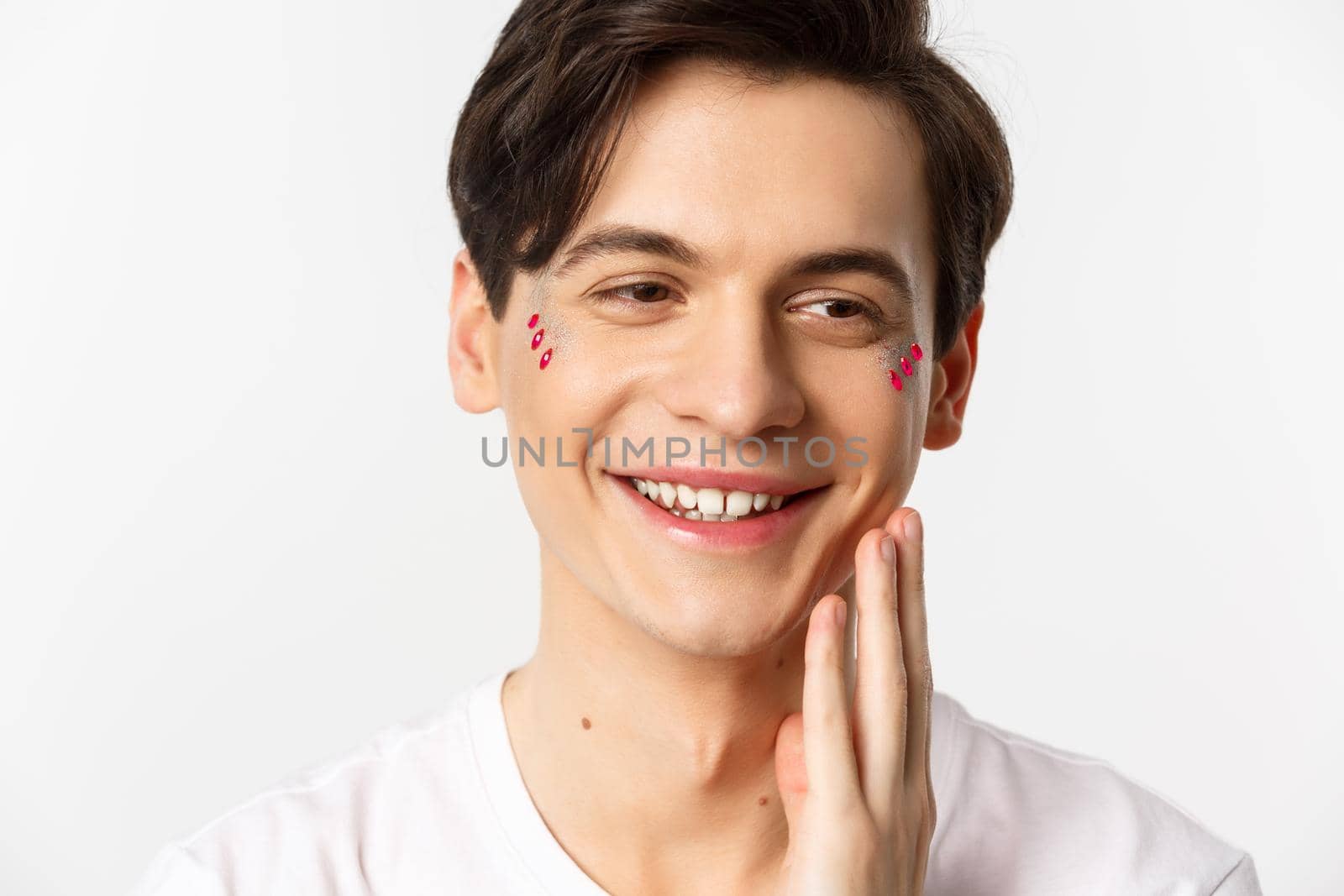 People, lgbtq and beauty concept. Headshot of beautiful gay man with glitter on face, smiling and looking happy, touching cheek after kiss, white background.