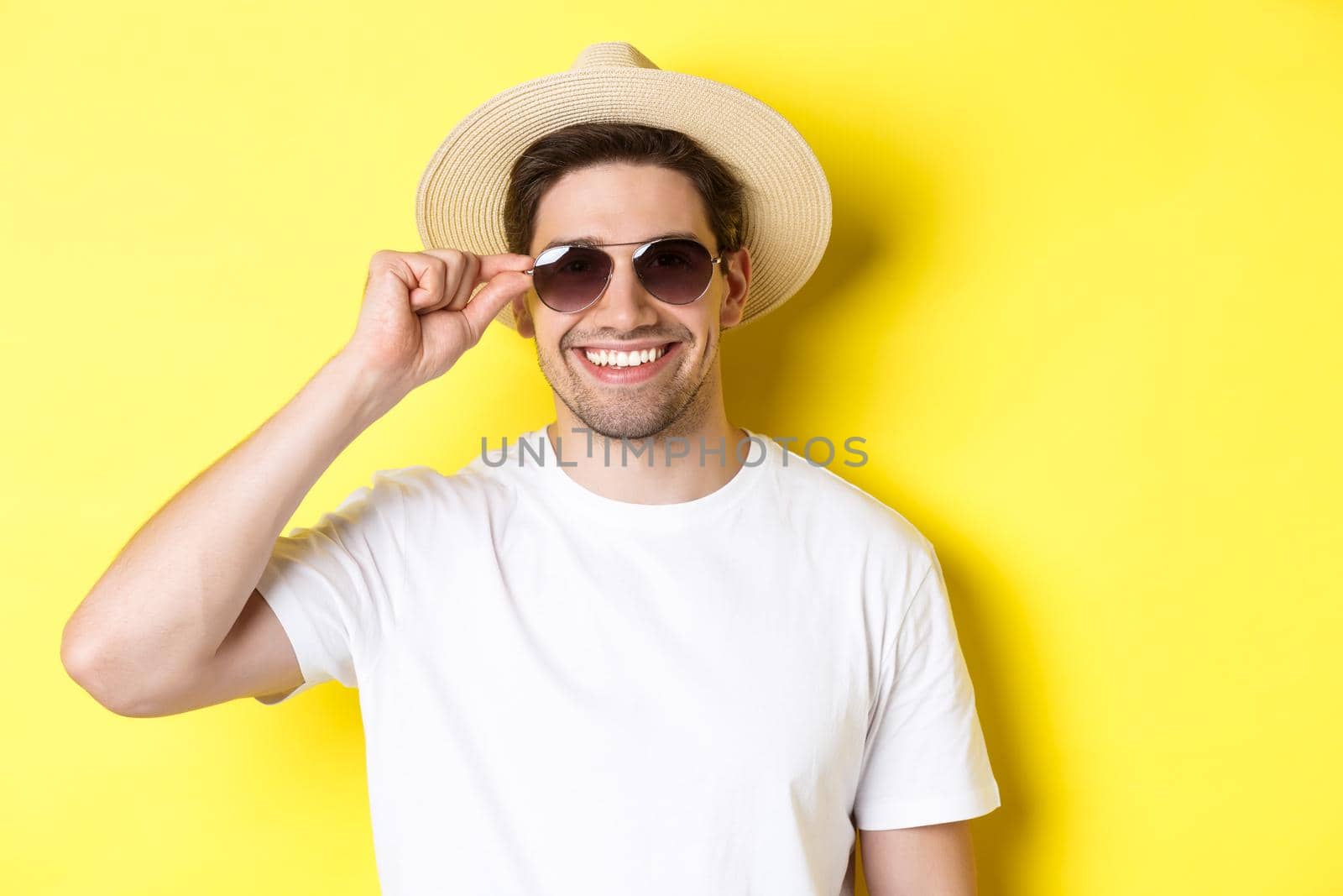 Concept of tourism and vacation. Close-up of handsome man tourist looking happy, wearing sunglasses and summer hat, standing over yellow background by Benzoix