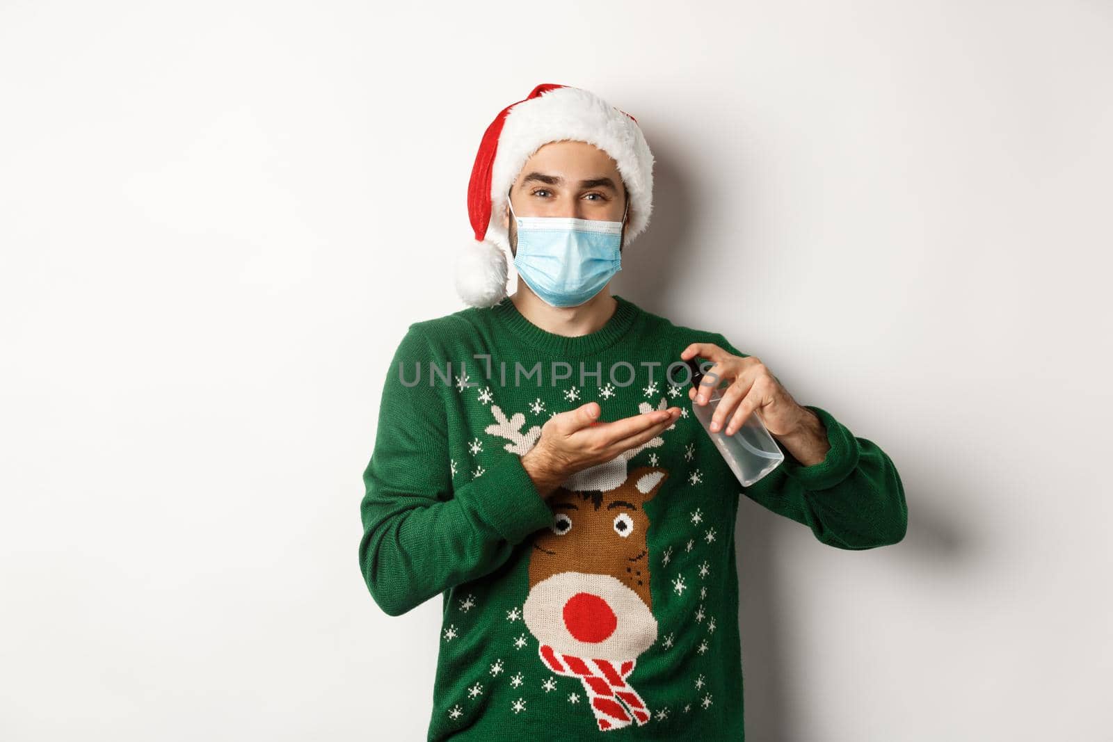Concept of covid-19 and Christmas holidays. Caucasian man in face mask and sweater using antiseptic, clean hands with sanitizer, standing over white background.