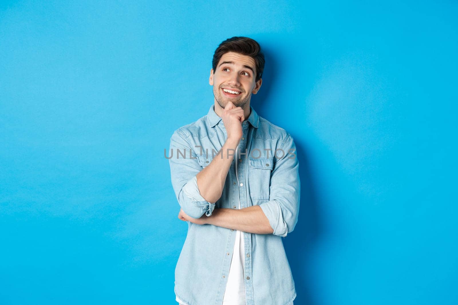 Portrait of thoughtful handsome man with beard, standing in casual outfit, looking at upper left corner and smiling, imaging or dreaming about something, standing over blue background by Benzoix