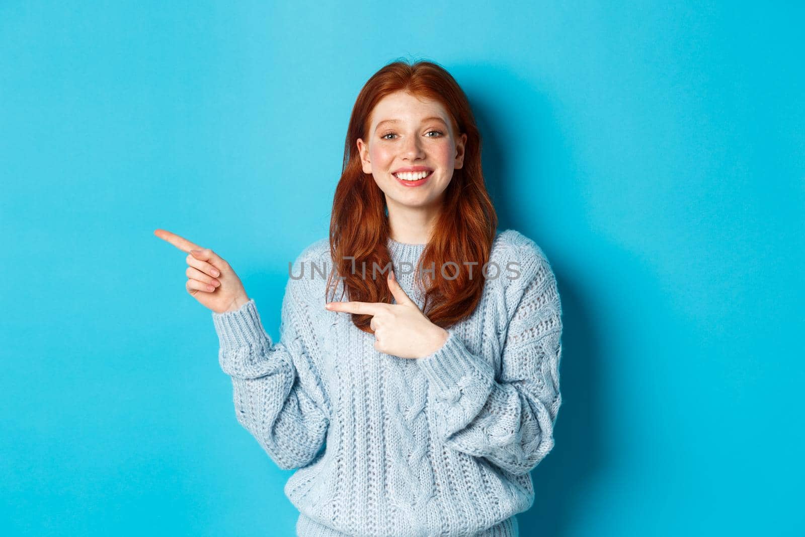 Cute teenager girl with red hair and freckles, pointing fingers left at logo and smiling, showing advertisement, standing over blue background.