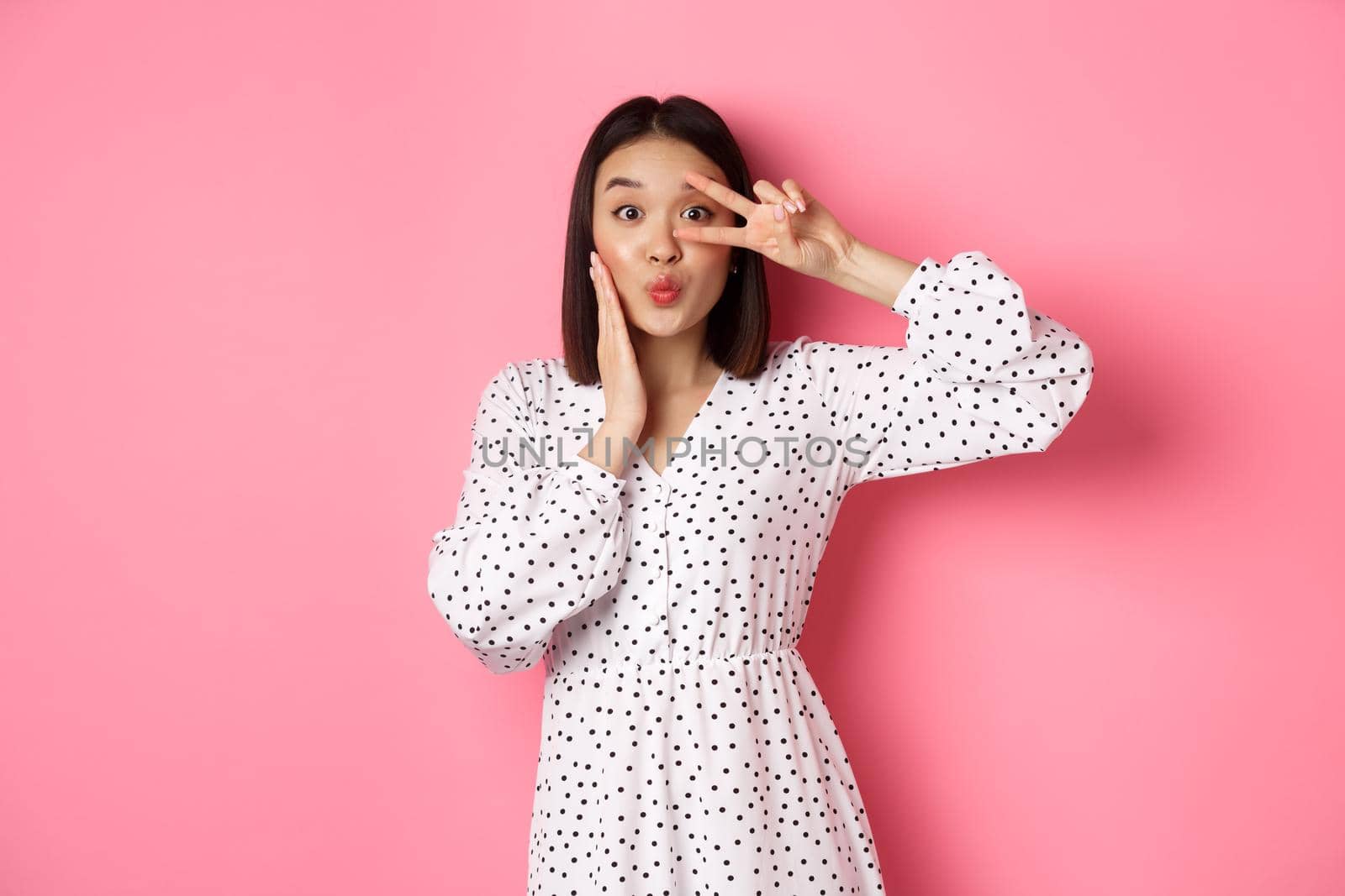 Coquettish asian woman in dress showing peace sign on eye, pucker lips for kiss, standing silly against pink background.