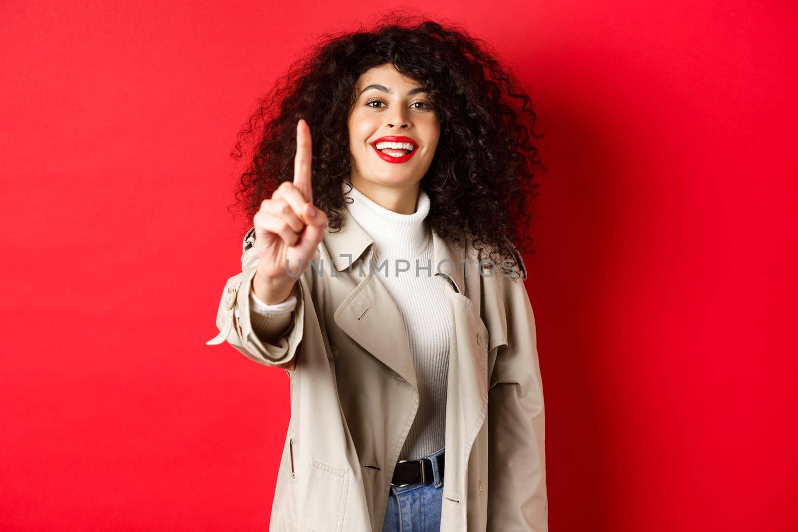 Cheerful woman in trench coat, showing number one finger and smiling, standing on red background.