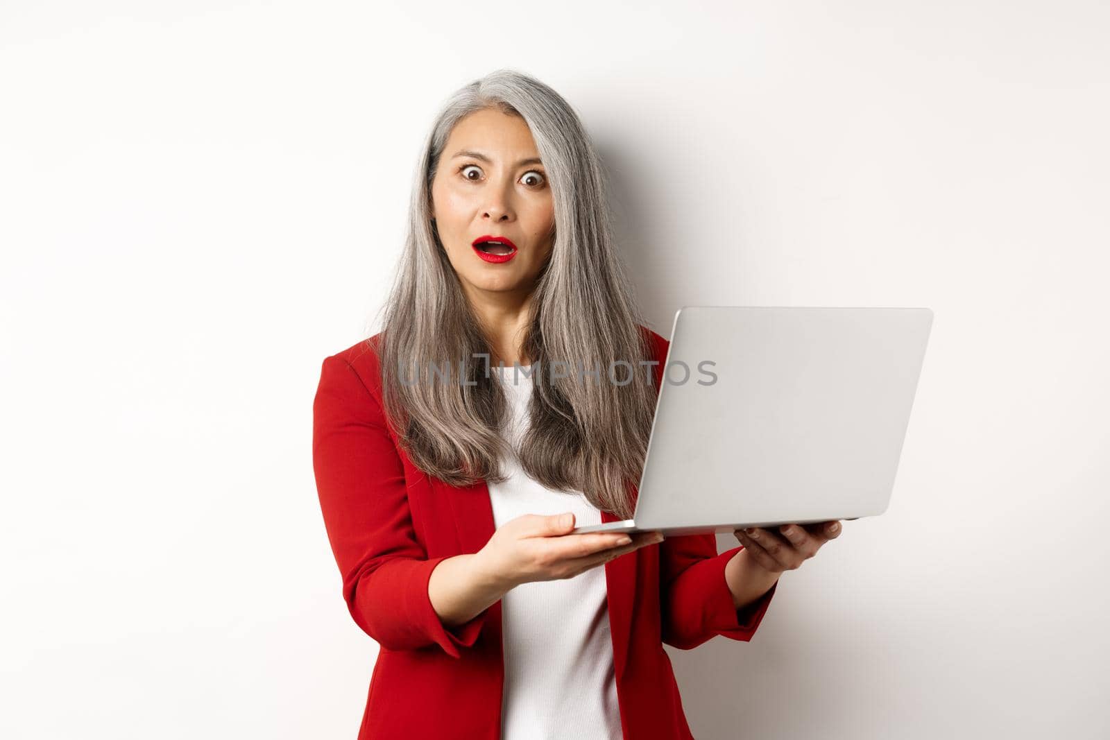 Business. Impressed asian old woman working on laptop, staring at camera surprised, standing over white background.