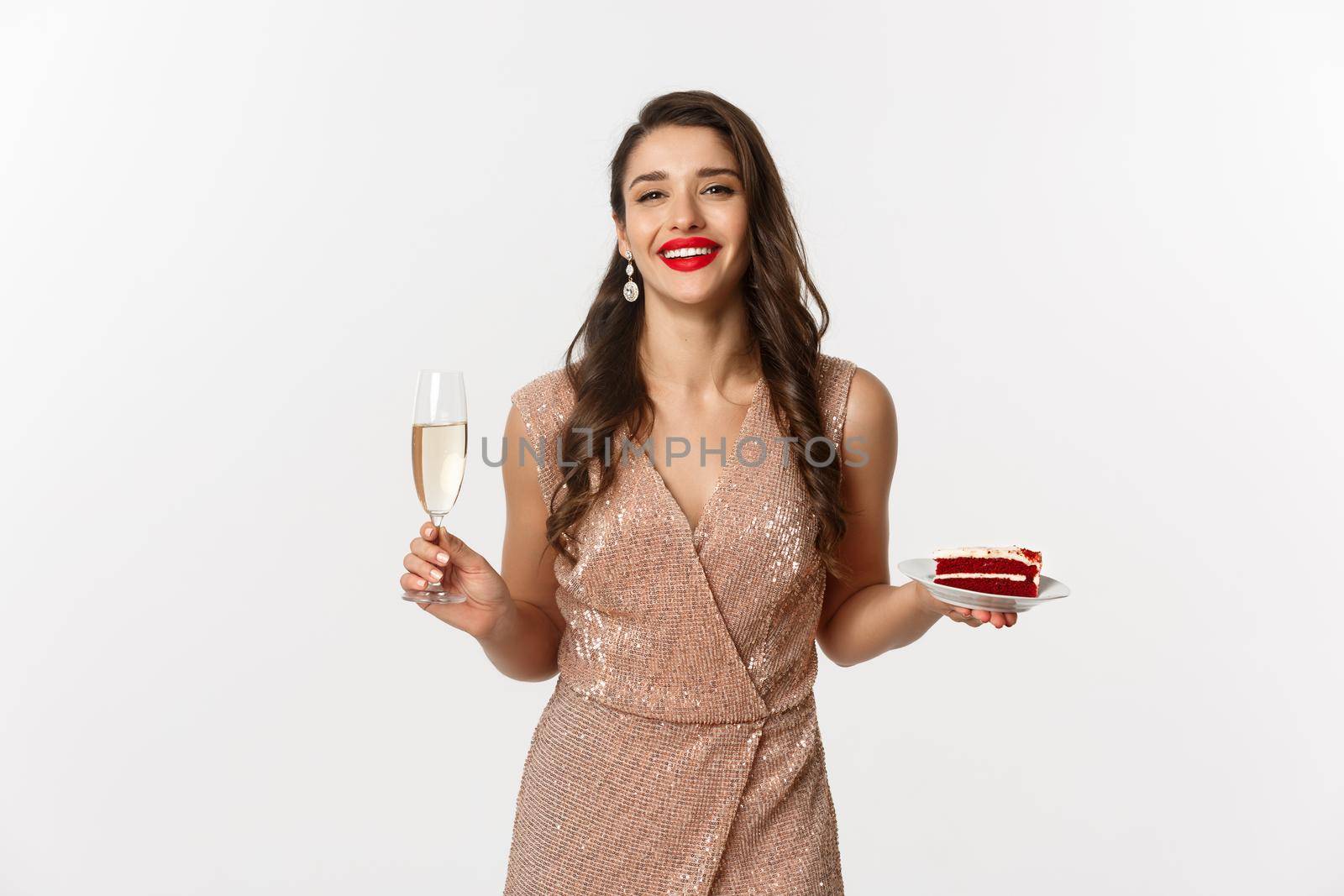 Party and celebration concept. Elegant woman with red lips, glamour dress, drinking champagne and eating cake, standing over white background.