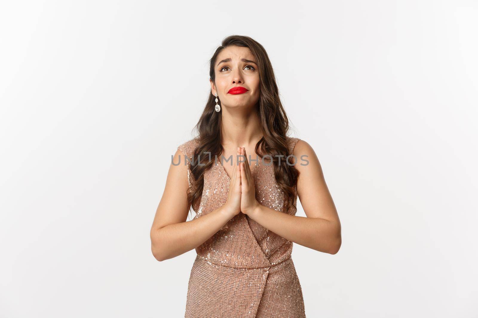 Celebration and party concept. Hopeful young woman begging for help, looking up and praying, pleading God, standing in glamour dress over white background.