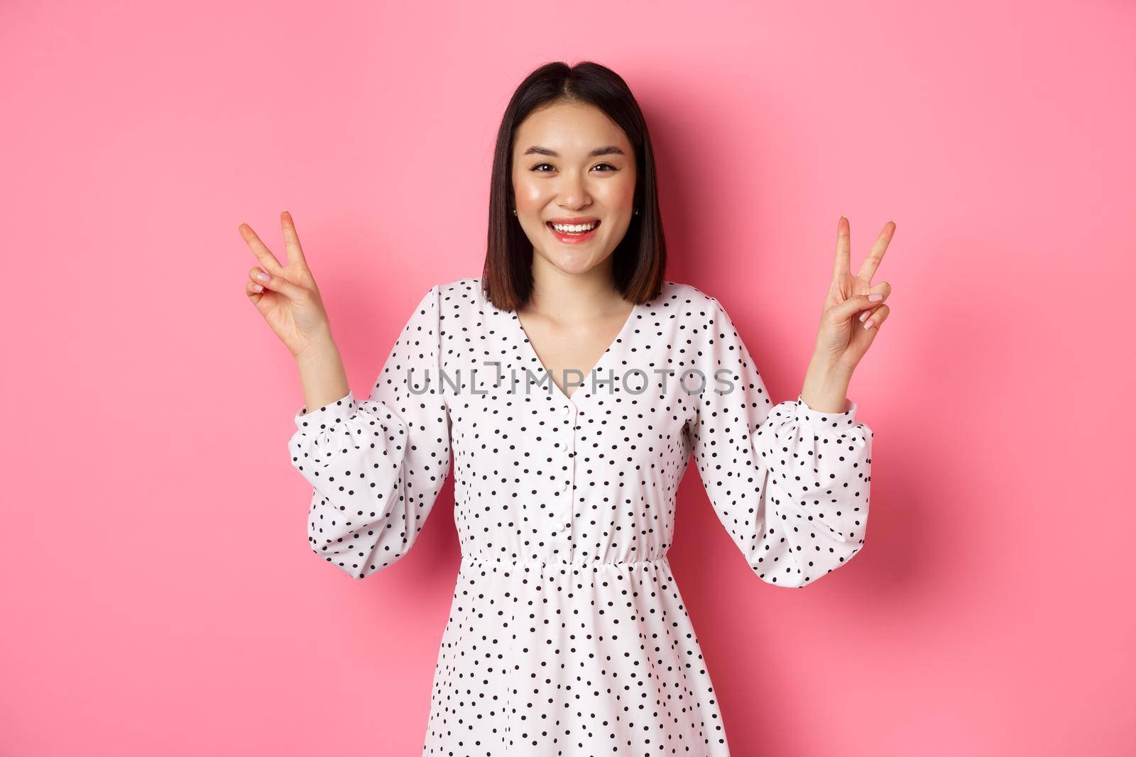 Cute asian brunette girl in dress smiling, showing kawaii peace signs and looking happy, standing over pink background.