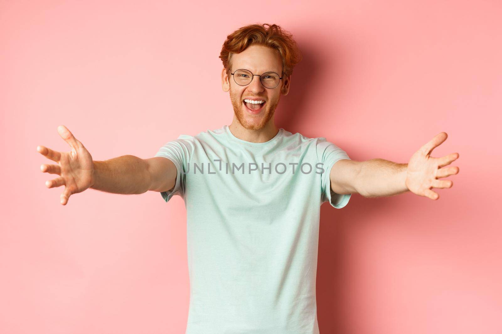 Young friendly man with red hair and beard reaching hands for hug, stretch out arm in warm welcome, smiling happily, standing over pink background.