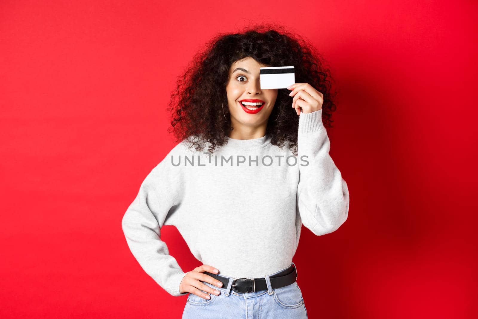 Stylish girl with curly hair showing plastic credit card over eye and smiling, standing against red background.