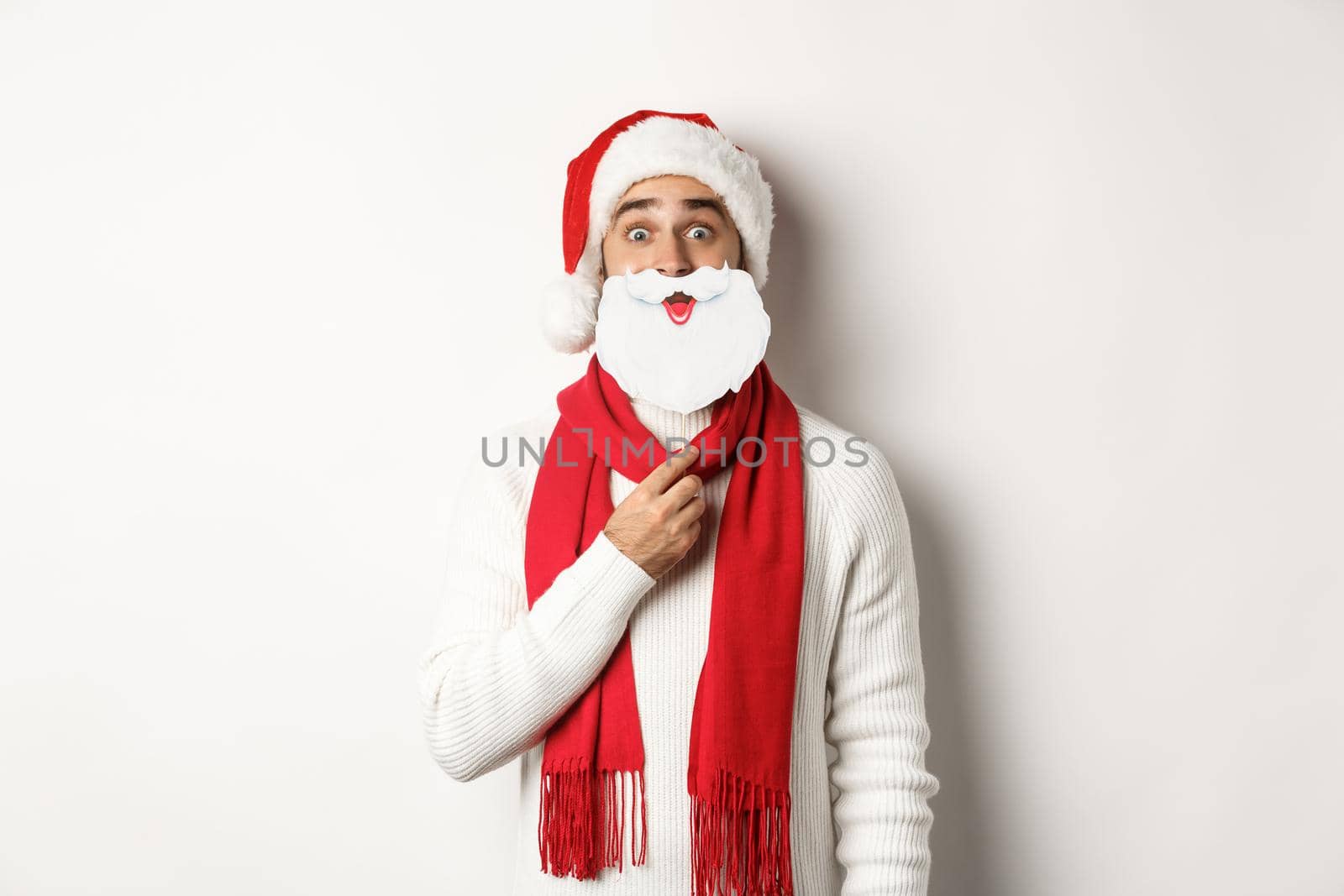 Christmas party and celebration concept. Funny young man in Santa hat holding white beard mask and making faces, enjoying New Year, white background.