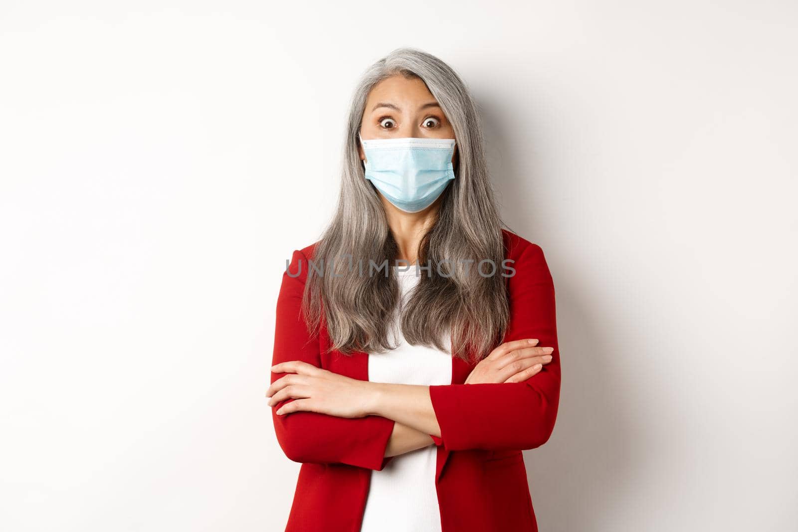 Covid-19 and business people concept. Asian businesswoman in face mask looking surprised, staring at camera in awe, standing over white background.