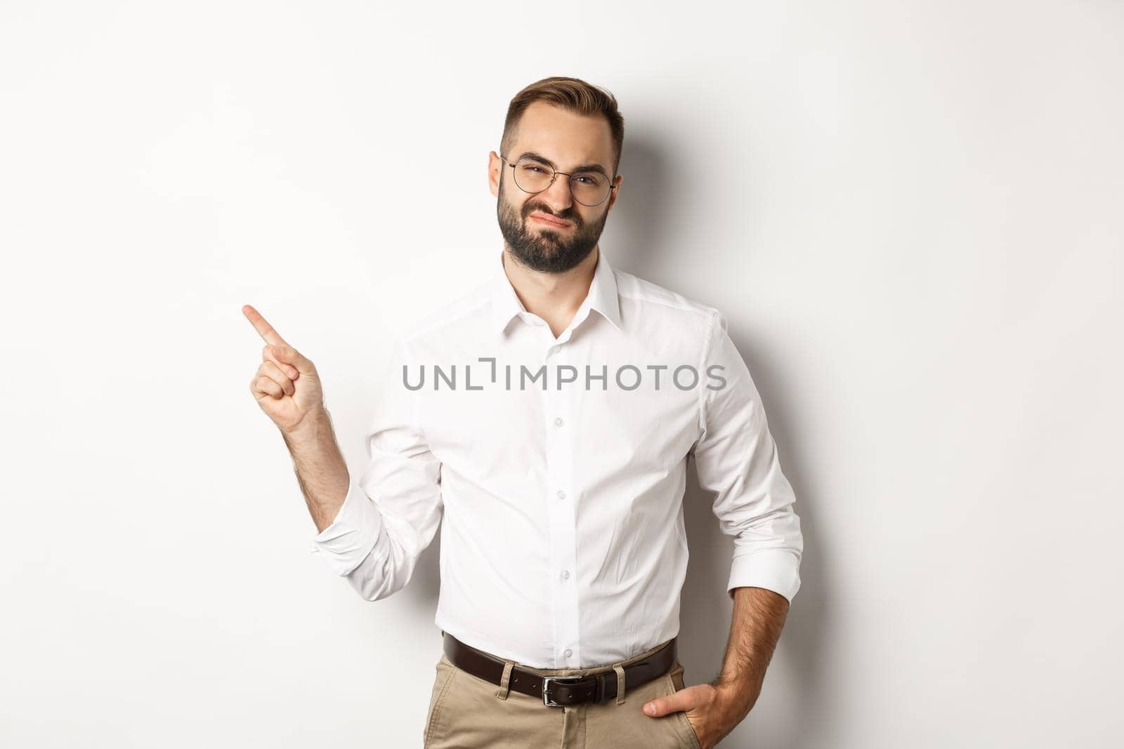 Skeptical male entrepreneur grimacing, pointing finger left with displeased face, complaining on product, white background.