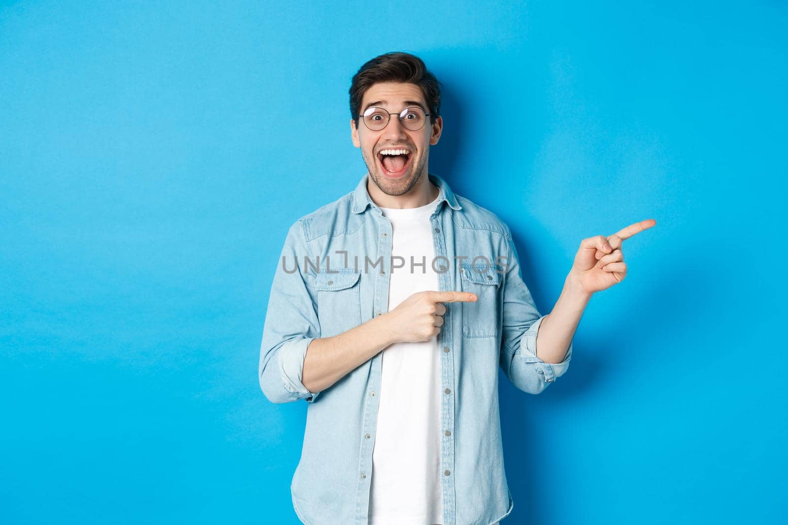 Surprised man in glasses pointing right at copy space, showing promo offer on blue background, standing over blue background.