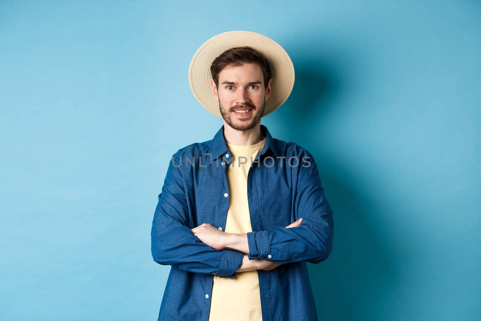 Portrait of cheerful guy going on vacation in straw summer hat, cross arms on chest and smiling, looking confident, standing on blue background.