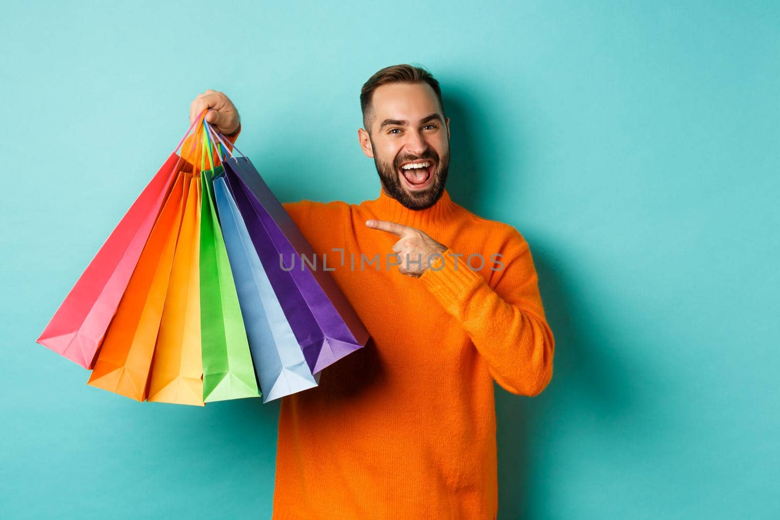Happy handsome man holding shopping bags and smiling, pointing at purchased items and recommending store, standing over turquoise background by Benzoix