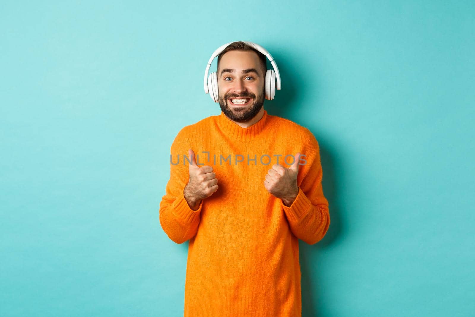 Amazed adult man listening music in headphones, looking at camera impressed and showing thumbs-up in approval, recommending, standing over turquoise background.