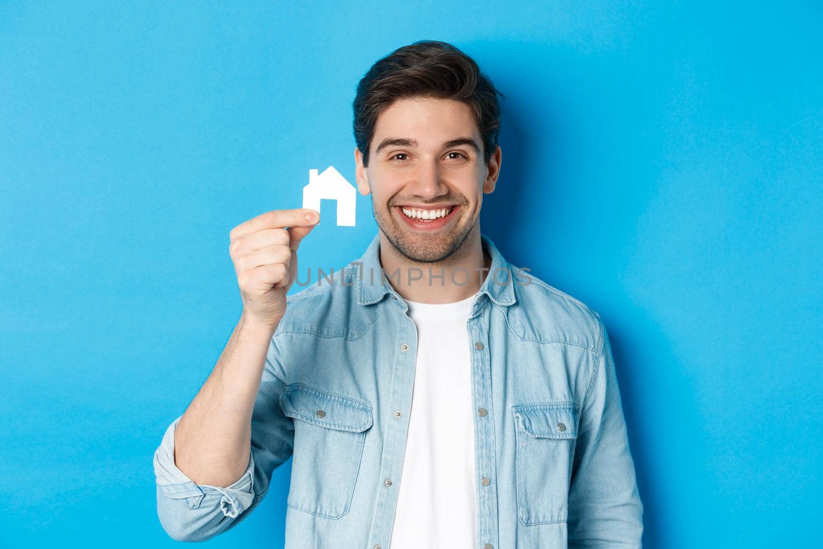 Real estate concept. Young handsome man smiling, showing small paper house mockup, searching for apartment, standing over blue background.