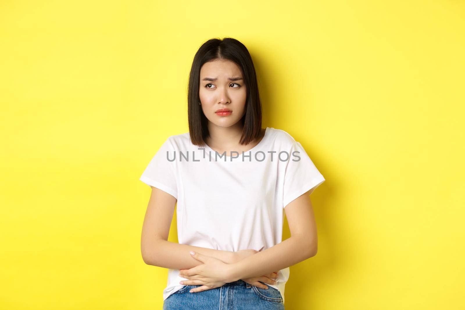 Image of asian woman feeling sick, having painful cramps, holding hands on belly and frowning from pain, discomfort during menstrual period, standing over yellow background.