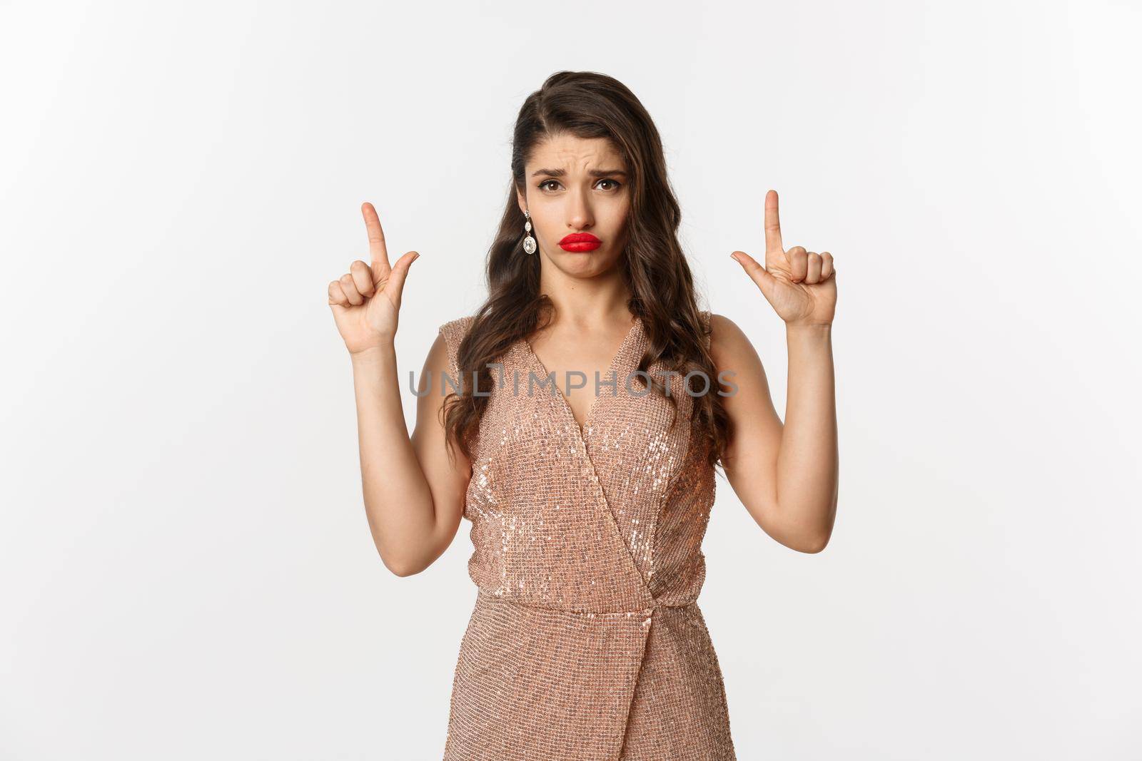 New Year, christmas and celebration concept. Gloomy and sad glamour girl in party dress pointing fingers up, sulking and crying about something, standing over white background.
