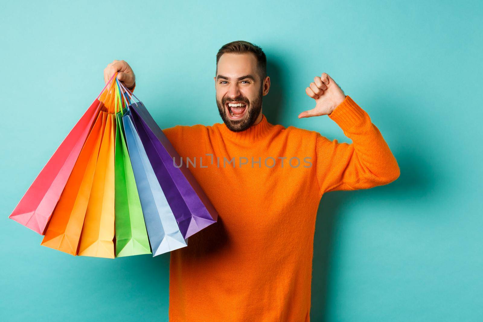 Handsome bearded man in orange sweater, buying gifts, pointing at himself and showing shopping bags, standing over blue background by Benzoix